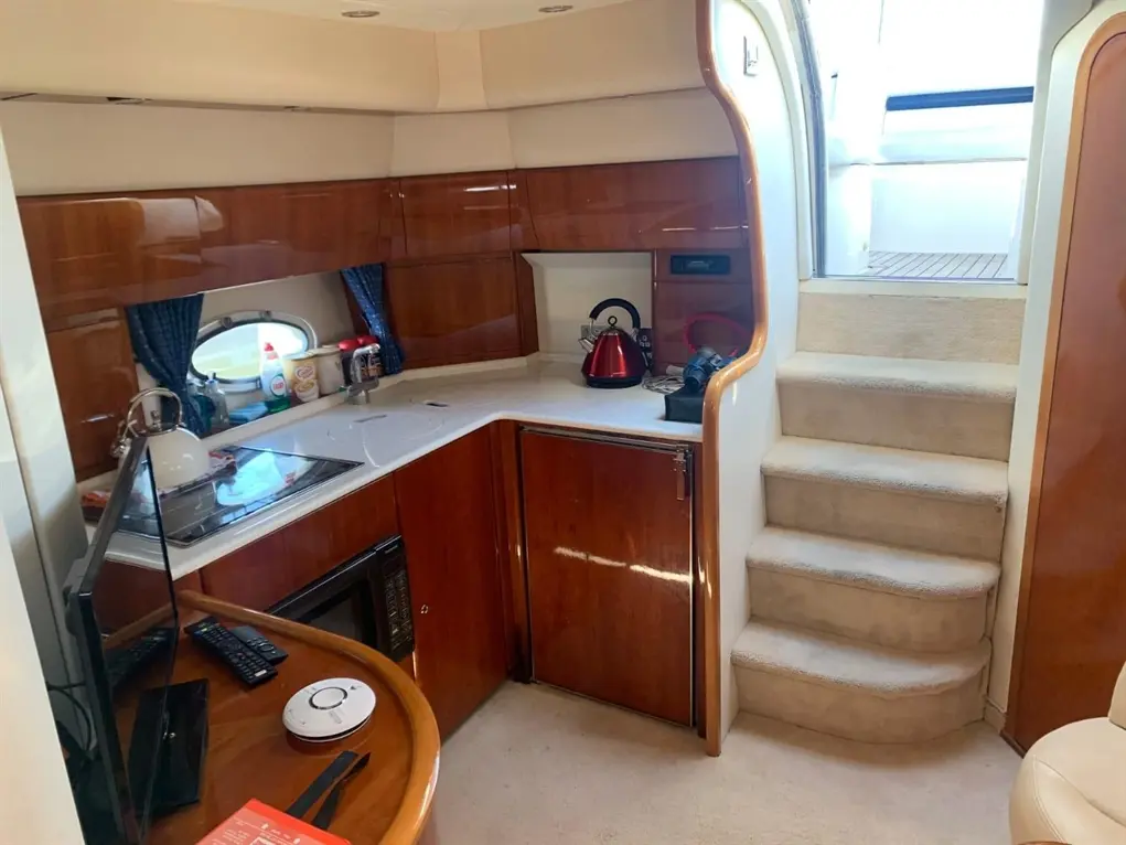 Modern boat interior featuring a kitchenette, kettle, TV, and stairs leading to the deck, with blue curtains and wooden cabinetry.